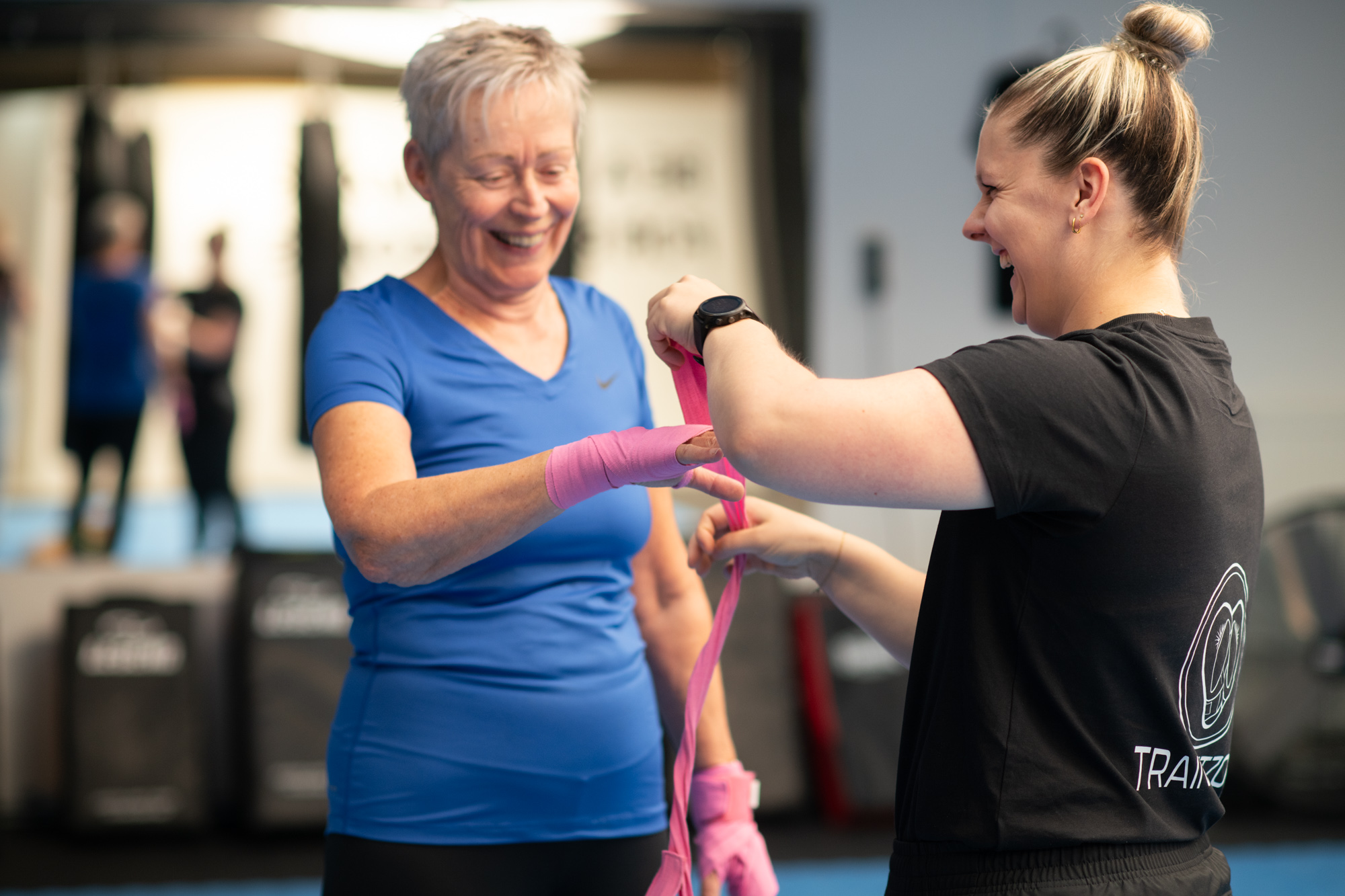 Esmee helpt een deelnemer met boksbandages; beiden lachen tijdens de voorbereiding op de training.